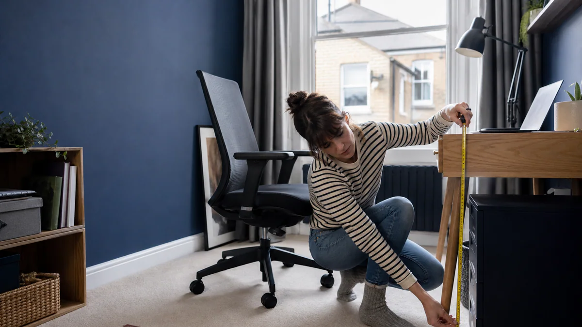 Person measuring desk and knee clearance beside an adjustable chair in a compact home office.